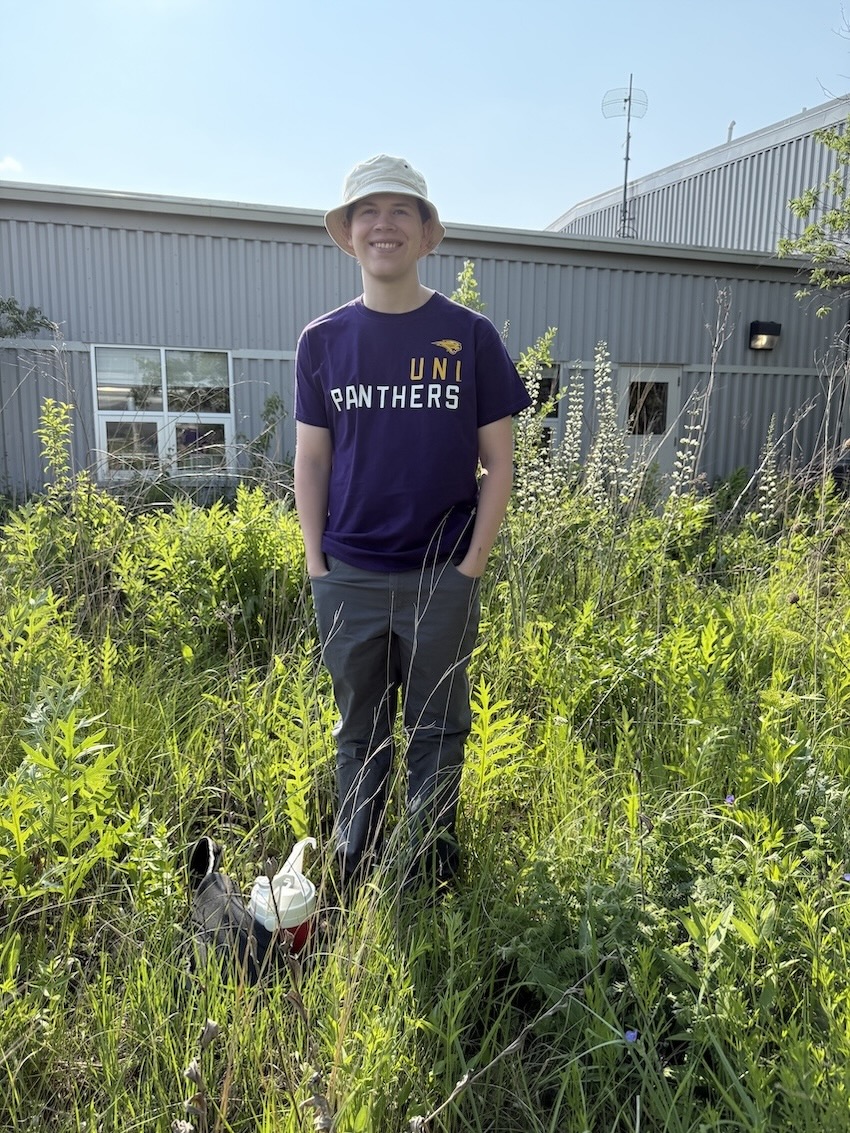Ethan Hughes wearing purple UNI Panthers t-shirt, a bucket hat, and standing in prairie plants outside of Tallgrass Prairie Center building
