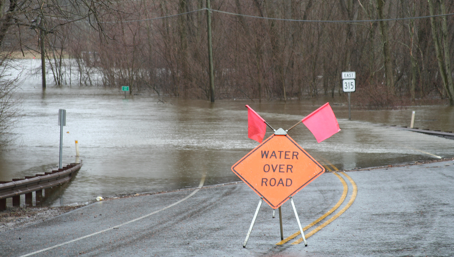 An orange sign reading "Water Over Road" is positioned on a flooded rural road.