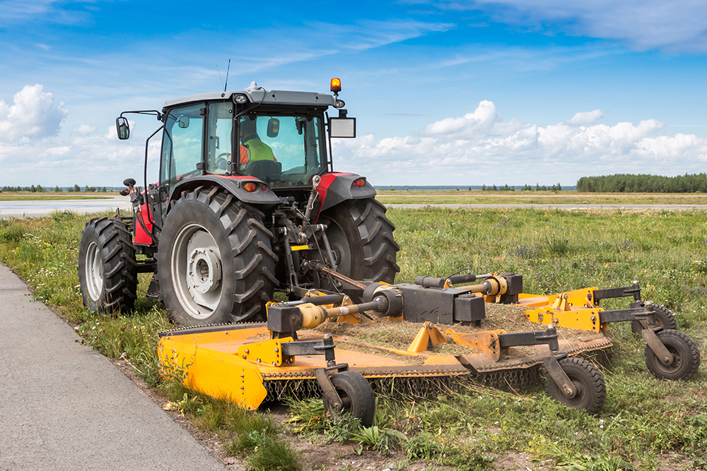 A tractor with large wheels pulls a mower through vegetation in the roadside.