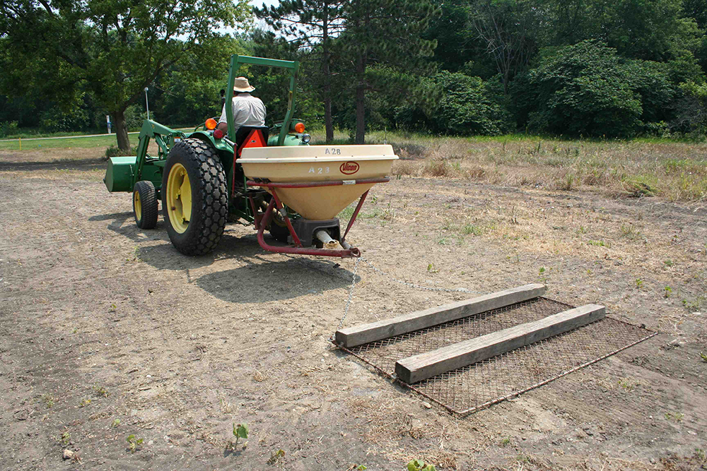 A person driving a green tractor pulling a white broadcast seeder.