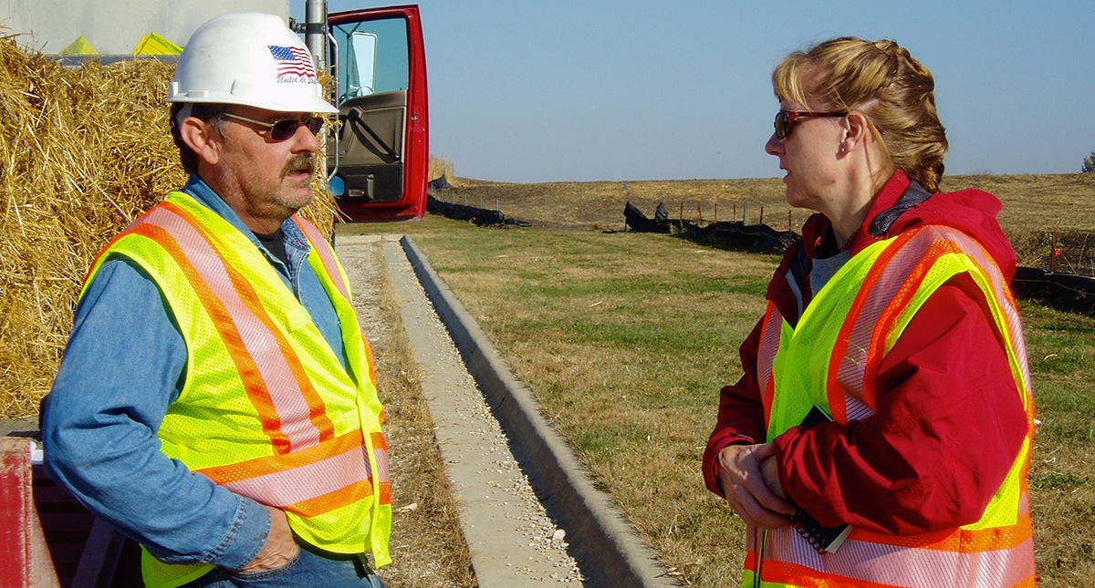 A man leaning on a load of hay in the back of a truck in a hard hat and safety vest chats with a woman in a safety vest. They stand on the side of a road