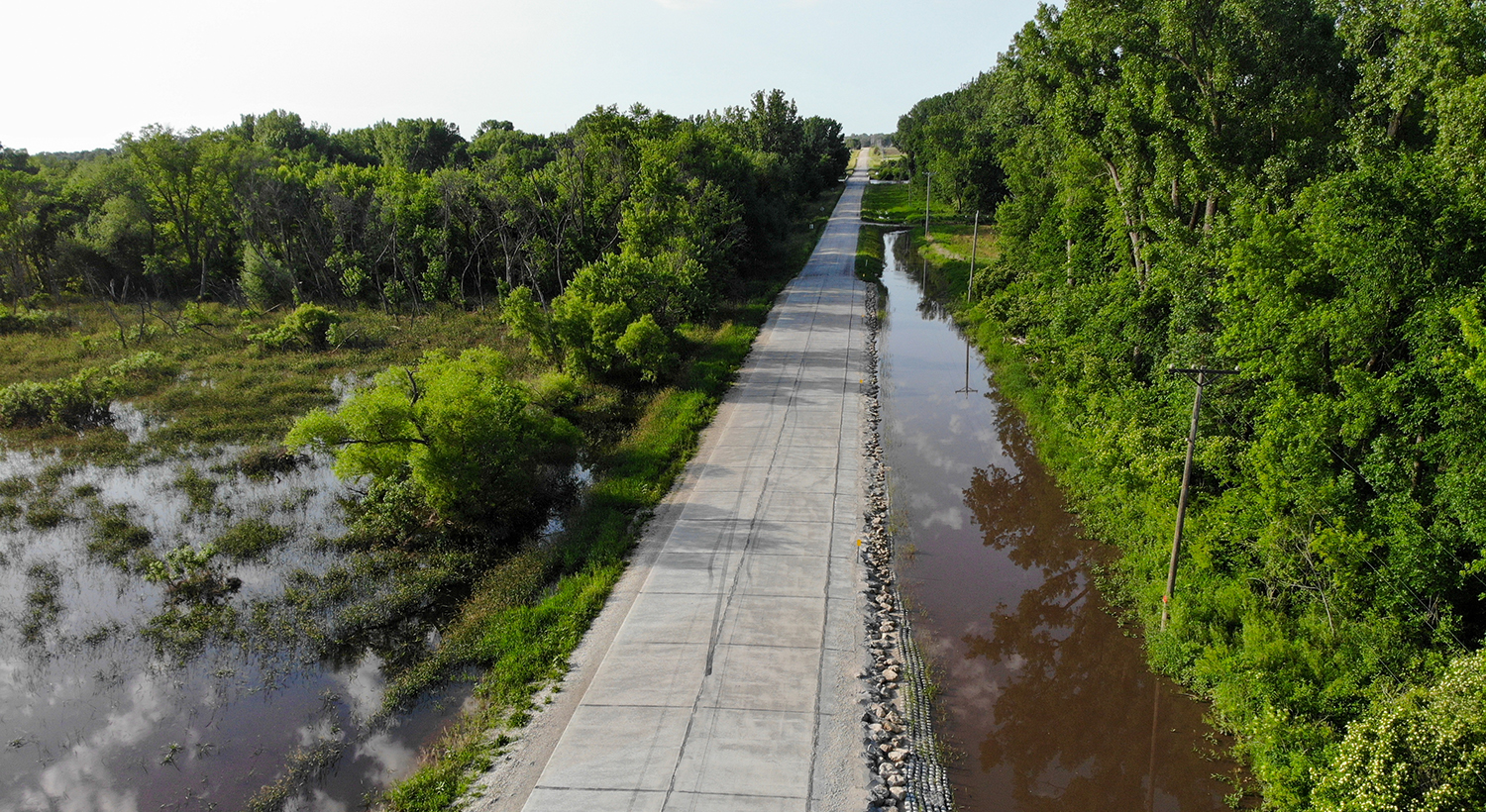 A road through the middle of a flooded forested area, as erosion control measures on both sides keep the road from eroding and flooding.