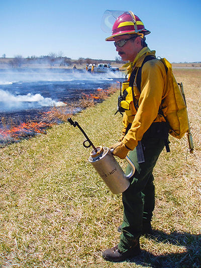 A man is holding a drip torch and dressed in controlled burn gear standing next to a controlled burn of roadside vegetation.