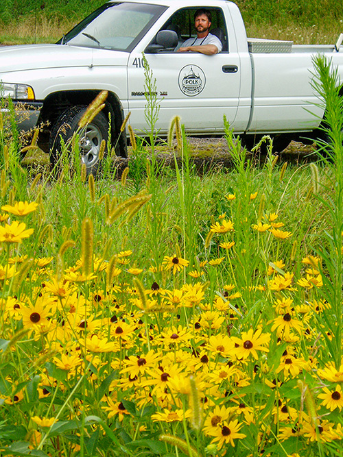 A roadside manager sits in a pickup truck next to roadside vegetation featuring yellow flowers.