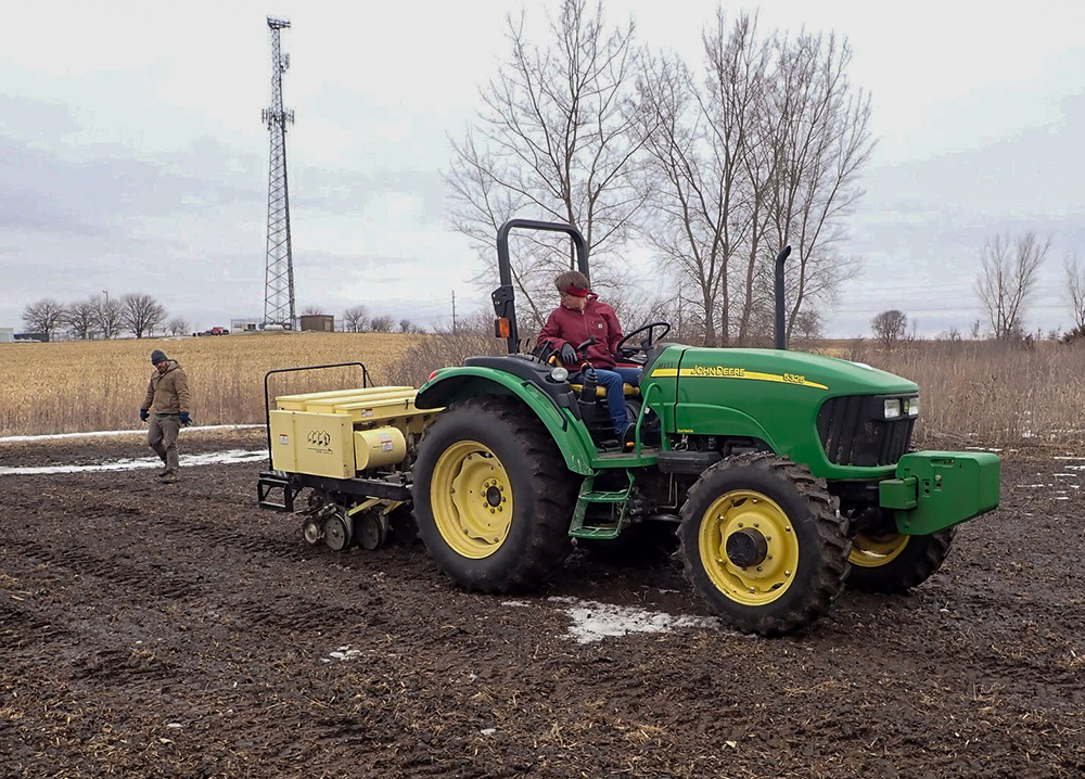 A green tractor trailing a seeder traverses a field in winter.