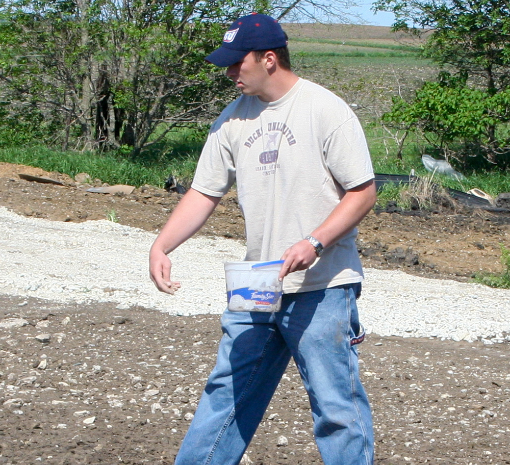 A person in a white shirt holds a small bucket of seeds and scatters prairie seed by hand.