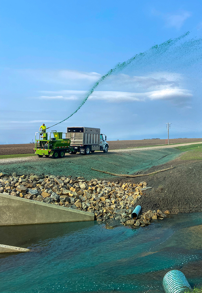 A person sprays hydromulch from a piece of equipment being towed behind a box truck into roadsides near a creek.