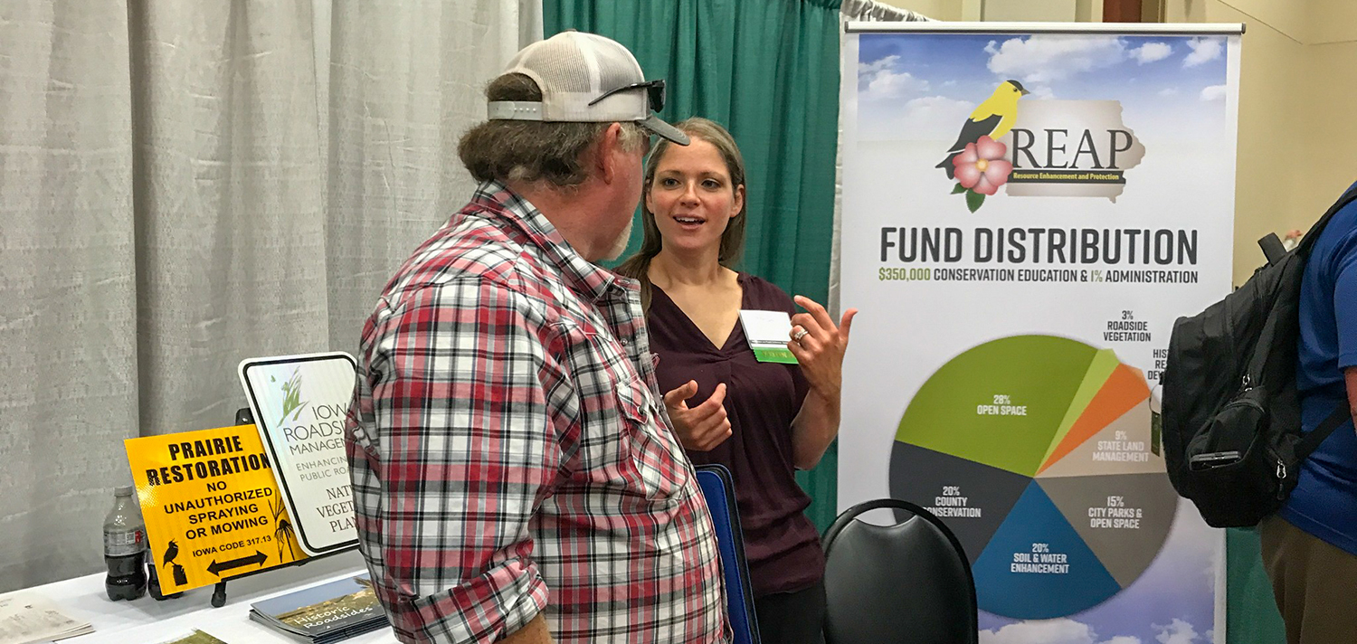 Linn County Roadside Manager Megan Huck speaks with an attendee at a conference with a poster for REAP fund distribution behind them.