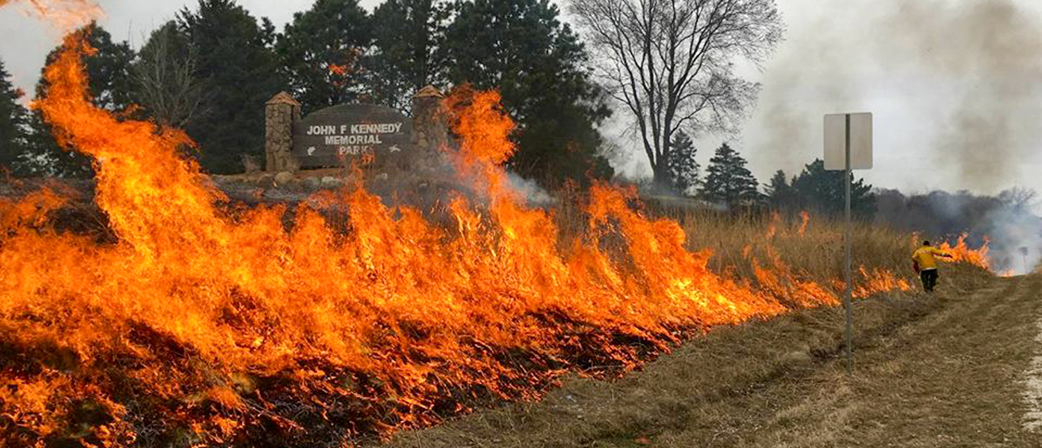 A prescribed fire burns on the side of the road as a staffer is seen in the background. A sign in the background reads "JOHN F KENNEDY MEMORIAL PARK"