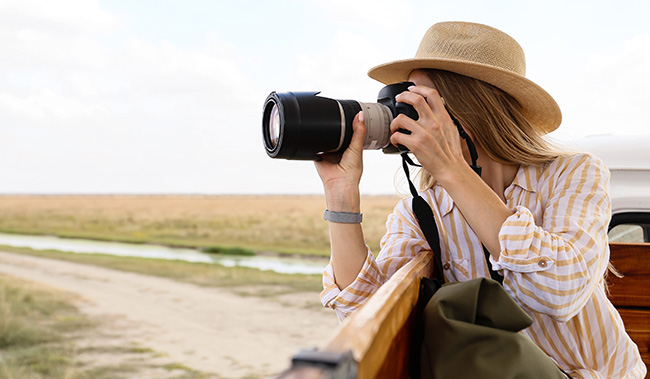 A woman wearing a hat holds a camera to take a photo from the back of a truck on the side of a rural road.