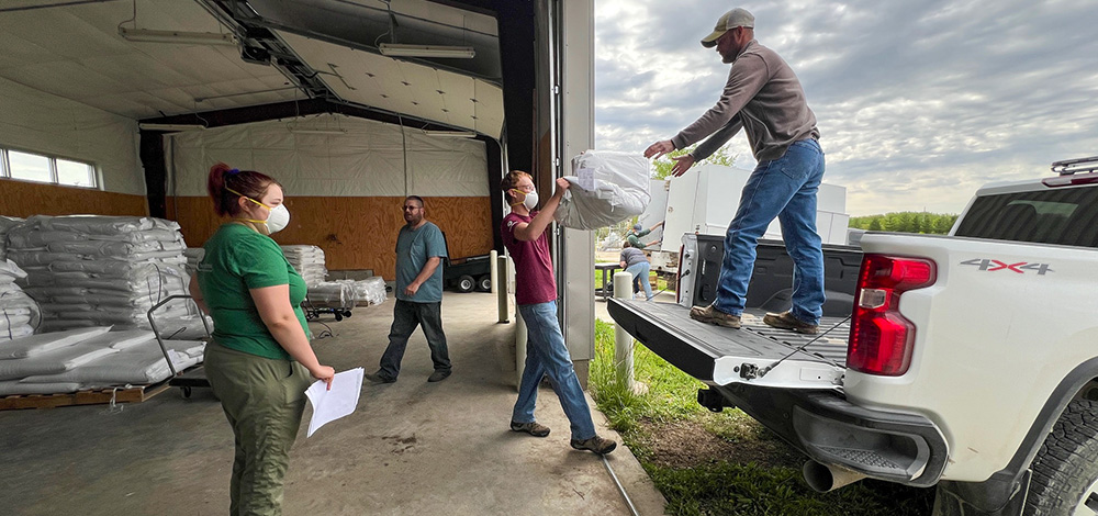 A man lifts a bag of seeds up to another man standing in the flatbed of a pickup truck as two other people look on.