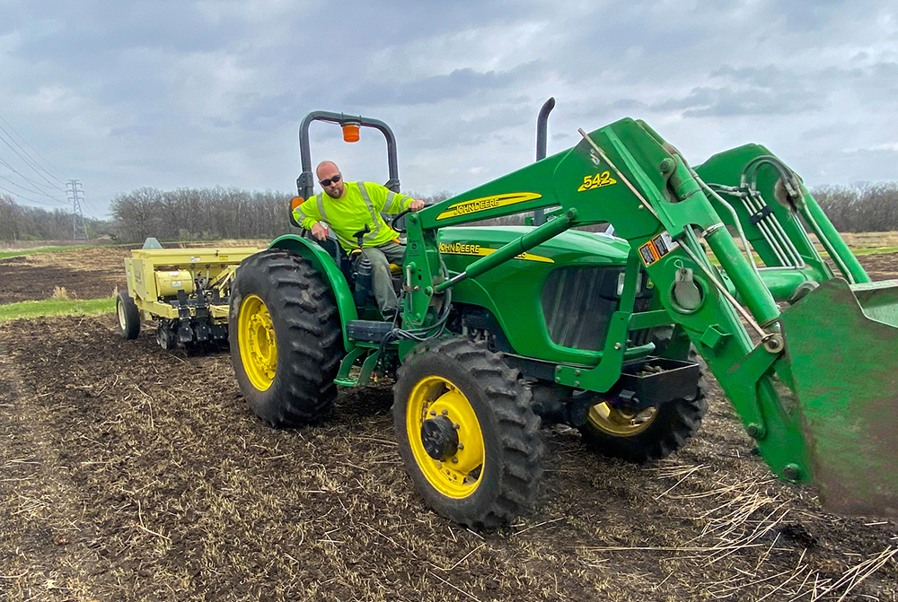 A person drives a green tractor pulling a yellow seeder over a bare roadside field.