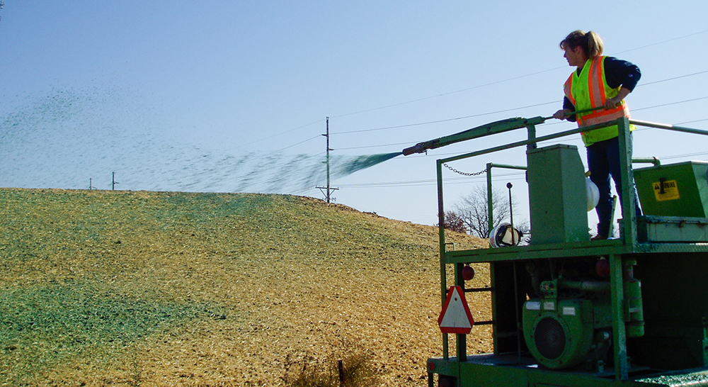 A person stands on top of a hydroseeder and sprays green slurry onto a sloped roadside.