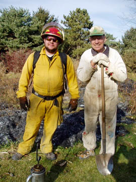 Two roadside staff members stand together in fire protection gear after a prescribed burn.