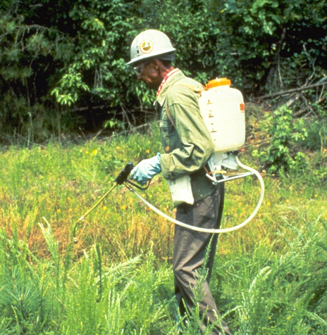 A worker in protective gear, including a helmet, gloves, and goggles, uses a backpack sprayer to spot-spray weeds.