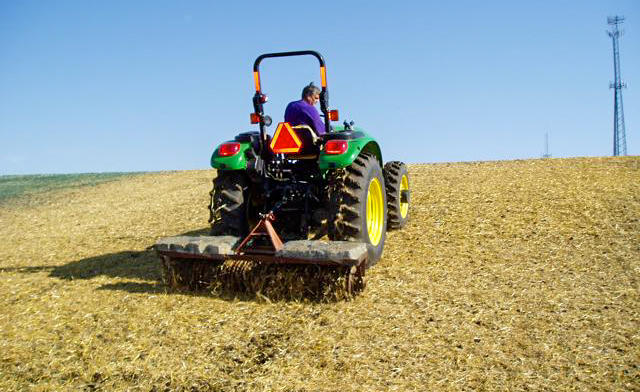 A person drives a tractor up a hill with a piece of equipment trailing behind while applying straw.