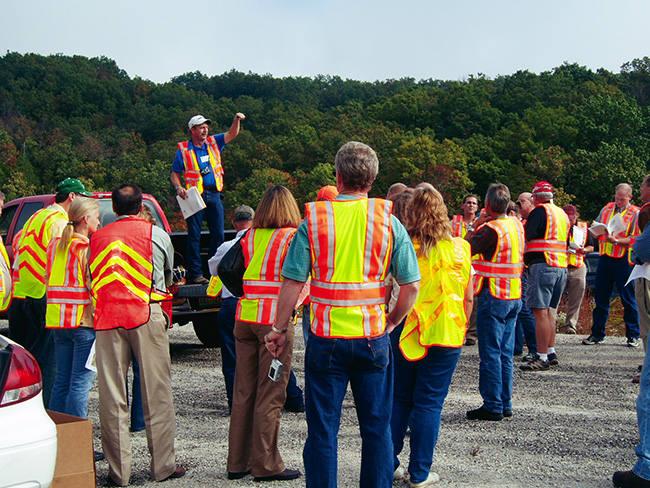 A group of people in safety vests listening to a person standing on the flatbed of a truck.