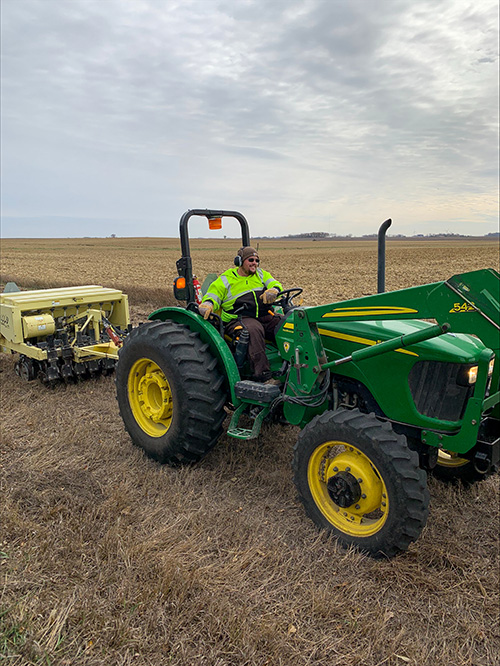 A man in a safety vest sits in a tractor pulling a native seed drill through a field.