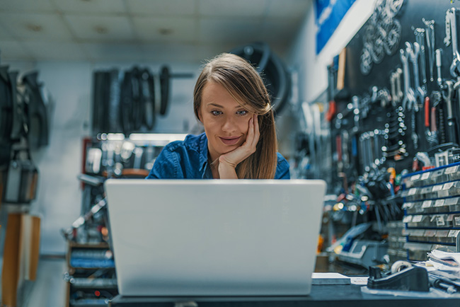 A woman rests her head on her palm and reads an email on her laptop in garage or equipment shop.