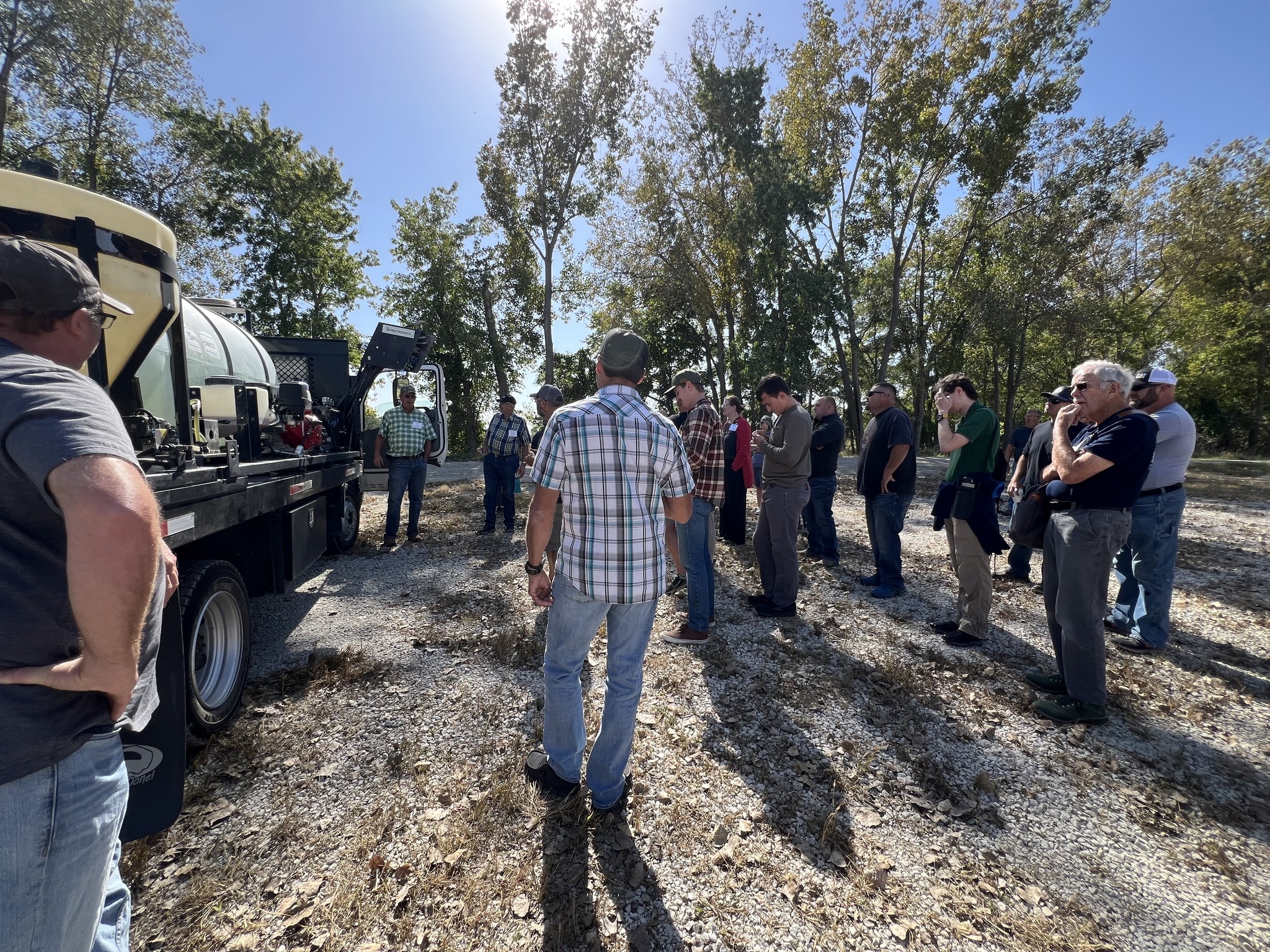 A group of roadside conference attendees on a sunny fall day standing outside listening to someone speak about a hydroseeder